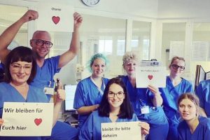 Pflegekräfte der Charité in Berlin in blauen Kitteln mit Postern "bleibt ihr bitte daheim" , "wie bleinen für Euch hier" und "wir halten durch"